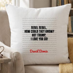 May include: A white square cushion featuring the lyrics "REBEL REBEL, HOW COULD THEY KNOW? HOT TRAMP, I LOVE YOU SO!" and the name "David Bowie". The cushion is on a beige armchair, with a wooden side table and a coffee cup.