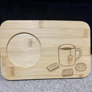 May include: A rectangular bamboo tea tray with a circular recess for a mug and etched illustrations. The tray features a mug with a teabag, and three biscuits: a Bourbon, a Custard Cream, and a heart-shaped biscuit.