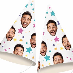 May include: Two white party hats featuring a repeating pattern of a smiling man's face and colourful stars in shades of pink, yellow, teal, and purple. Each conical hat includes a thin white elastic chin strap for securing the headwear in place.