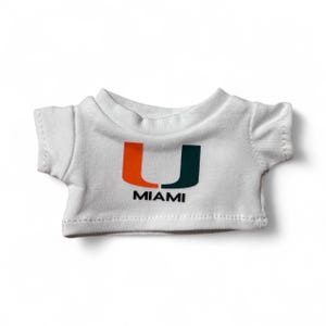 May include: A small white t-shirt with the word "MIAMI" printed in black below a stylized orange and green "U" logo. The shirt is made of a soft fabric and has short sleeves.