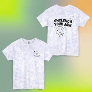 Pode incluir: Duas camisetas brancas com um padrão tie-dye. Uma camisa tem um pequeno gráfico de rosto sorridente na frente. A outra camisa tem o texto "UNCLENCH YOUR JAW" acima de um gráfico de rosto sorridente derretendo nas costas.