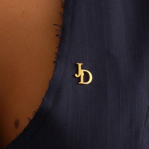 May include: A gold-colored pin featuring the initials "JD" in a stylized script. The pin is affixed to a dark navy blue garment, creating a contrast between the bright metal and the fabric. The pin is a small, elegant accessory.
