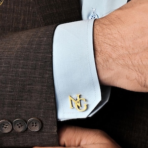 May include: Gold-colored cufflinks with the initials "NG" are displayed on a light blue dress shirt. The cufflinks are worn with a brown suit jacket. The jacket has four dark buttons.