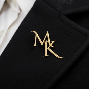 May include: A gold-colored pin featuring the intertwined letters "MK" is affixed to a black coat. The pin is a stylized monogram, with the letters elegantly curved and connected. The coat is worn over a white collared shirt.