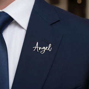 Könnte beinhalten: Ein silberfarbener Anstecker mit dem Wort "Angel" in Schreibschrift ist an einem marineblauen Anzugsakko befestigt. Der Anstecker befindet sich in der Nähe des Revers. Ein weißes Hemd und eine blaue Krawatte sind ebenfalls zu sehen.