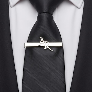 Op de afbeelding: Een zilveren stropdas met de initialen "AR" in een gestileerd lettertype wordt getoond op een zwarte stropdas. De stropdas maakt deel uit van een formele outfit, inclusief een wit overhemd en een zwart colbert.