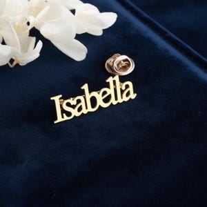 Könnte beinhalten: Ein goldfarbener Ansteckpin mit dem Namen "Isabella" in Schreibschrift. Der Pin befindet sich auf einem dunkelblauen Samthintergrund, mit weißen Blumen. Der Pin hat einen Standard-Pin-Verschluss.