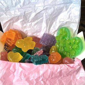 May include: A box filled with colorful, translucent candies in various shapes, including hearts, flowers, and geometric forms. The candies are orange, yellow, green, blue, and purple. The box has a pink interior and is lined with white tissue paper.