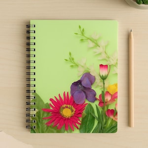 Puede incluir: Un cuaderno con espiral y cubierta verde claro, adornado con un vibrante arreglo floral. Las flores incluyen una gran margarita rosa, violetas moradas y flores blancas. Un lápiz de madera descansa junto al cuaderno.