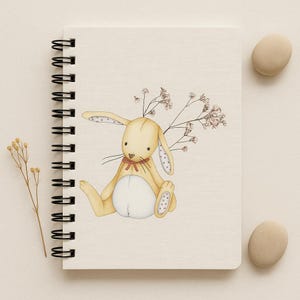 Puede incluir: Un cuaderno con espiral de color crema con una ilustración caprichosa de un conejito amarillo sentado con orejas con lunares rosas y una pajarita roja. El conejito está adornado con delicadas ramitas florales. Dos piedras lisas de color beige descansan junto al cuaderno.