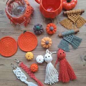 Può includere: Una collezione di decorazioni a tema autunnale che include un barattolo di vetro con copertura in macramè arancione, una zucca in ceramica arancione, due sottobicchieri all'uncinetto, piccole zucche all'uncinetto, due arazzi e due portachiavi.