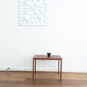 May include: A rectangular wooden side table with four straight legs and a smooth top surface, featuring a small black bowl centred on it, positioned below a white wall-mounted panel with a geometric, three-dimensional relief pattern.