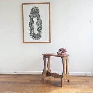May include: A wooden side table with curved legs and a rectangular top, featuring a vintage red rotary telephone placed on its surface, positioned below a framed black and white abstract line drawing of a figure in a wooden frame.
