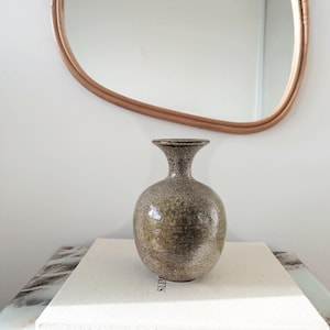 May include: A round ceramic vase with a narrow neck and a flared rim. The surface features a mottled, speckled texture in shades of earthy brown, tan, and grey, creating a variegated appearance across the entire rounded body of the vessel.