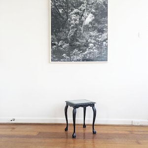 May include: A small black side table with a square top and four curved legs with ball and claw feet, positioned below a large rectangular framed black and white photograph of a dense forest scene hanging on a plain white wall.