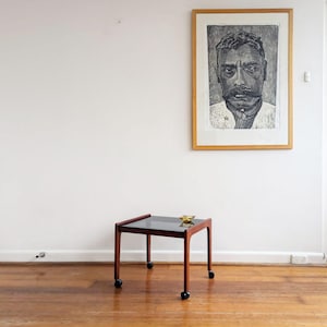 May include: A square side table with a dark wood frame and a black glass top, supported by four black casters. A small, gold-toned decorative bowl sits on the surface. Above the table, a framed black and white portrait print hangs on the wall.