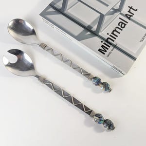 May include: A pair of silver-toned metal salad servers featuring a solid spoon and a slotted spoon. The handles are wrapped in wire with a zigzag pattern and finished with two decorative, marbled blue and grey beads at the end of each handle.