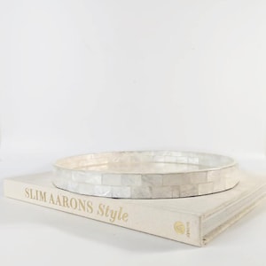 May include: A round tray featuring a mosaic of iridescent white mother-of-pearl tiles. The tray is positioned on top of a thick, cream-coloured hardcover book with the text Slim Aarons Style printed in gold on the spine and front cover.