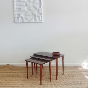 May include: A set of three nesting tables with dark brown tops and reddish-brown legs. A small, round, wooden bowl sits on the largest table. The tables are arranged in a stepped configuration. The smallest table is in front.