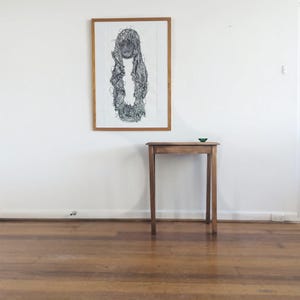 May include: A framed black and white artwork hangs on a white wall above a small wooden table. The artwork depicts a detailed, abstract figure. A small, green object sits on the table's surface. The room has a hardwood floor.