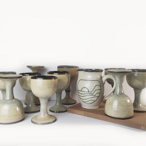 May include: A collection of beige ceramic goblets and a matching pitcher with a dark brown rim. The pitcher features a hand-drawn graphic of a sun setting over waves. Some goblets are inverted, and the set rests on a brown book labelled CHAGALL.