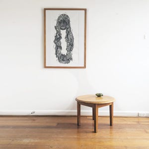 May include: A framed black and white ink drawing of a figure draped in intricate, textured lace-like fabric, displayed on a wall above a small, round, light-brown wooden table with three legs and a single small green object on its surface.