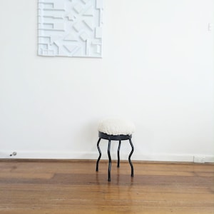 May include: A stool with a round, white, textured, fleece-like seat cushion and a black metal base featuring four thin, wavy, curved legs. Above the stool, a square white wall panel displays a geometric relief pattern of raised shapes.
