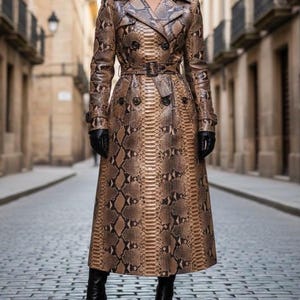 May include: A long, snakeskin-patterned trench coat in shades of brown and tan. The coat features a double-breasted design, a belt at the waist, and black gloves and boots. The coat is worn in a cobblestone street.