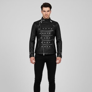 Könnte beinhalten: Schwarze Lederjacke mit hohem Kragen, asymmetrischem Reißverschluss und silberfarbenen Metallringen. Die Jacke hat ein militärisch inspiriertes Design mit dekorativen Reihen silberner Knöpfe und eine taillierte Silhouette. Das Model trägt eine schwarze Hose.