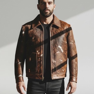May include: A brown leather jacket with a cowhide pattern, featuring a zipper closure and two front pockets. The jacket has a classic collar and cuffs. The jacket is worn over a black shirt and paired with dark wash jeans.