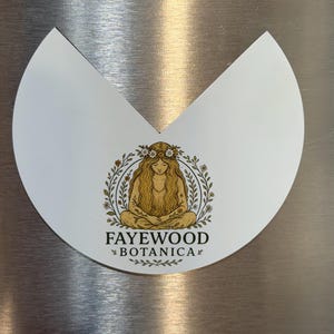May include: A white, semi-circular magnet with a V-shaped cutout at the top. It features a gold-toned illustration of a person with long hair and a flower crown, surrounded by a leafy wreath. The text FAYEWOOD BOTANICA is printed below.