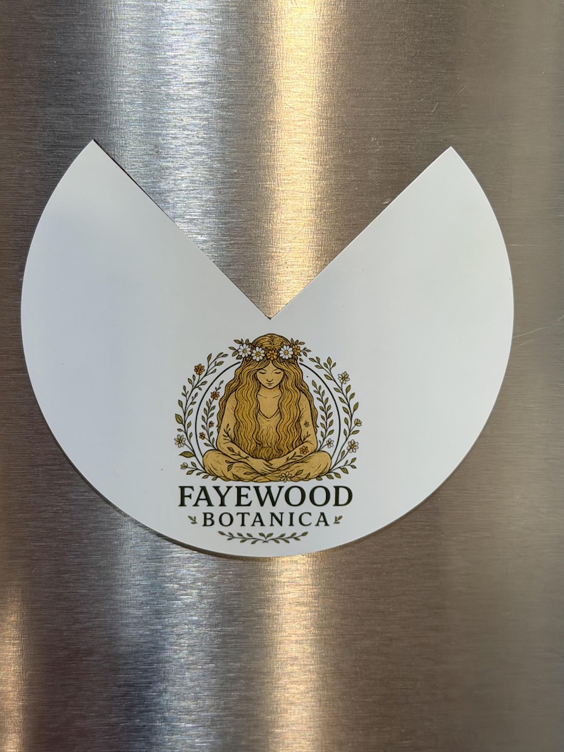 May include: A white, semi-circular magnet with a V-shaped cutout at the top. It features a gold-toned illustration of a person with long hair and a flower crown, surrounded by a leafy wreath. The text FAYEWOOD BOTANICA is printed below.