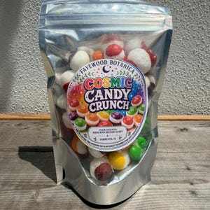 May include: A clear, resealable stand-up pouch filled with multicolored, freeze-dried candy. The label features the text Fayewood Botanica, Cosmic Candy Crunch, and Ingredients: Made with Skittles Candy, Sorrento, FL, with a colorful, starry design.