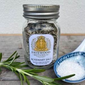 May include: A clear glass jar with a silver lid filled with rosemary salt. The jar has a label with the text "FAYEWOOD BOTANICA ROSEMARY SALT". Fresh rosemary sprigs and a small blue and white spoon with salt are next to the jar.