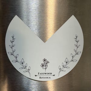 May include: A white, semi-circular sticker with a V-shaped cutout at the top. It features black line art of two leafy branches on the sides and a small floral illustration in the center above the text Fayewood Botanica.
