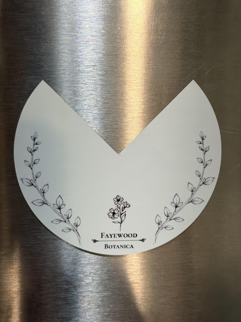 May include: A white, semi-circular sticker with a V-shaped cutout at the top. It features black line art of two leafy branches on the sides and a small floral illustration in the center above the text Fayewood Botanica.