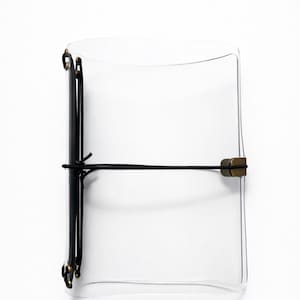 Pode incluir: Um diário retangular transparente com lombada preta e fecho elástico preto. O diário tem um fecho cor de latão e está sobre um fundo branco. O diário parece estar vazio.