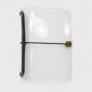 Puede incluir: Cubierta de cuaderno rectangular transparente con lomo negro y cierre elástico. La cubierta se asegura con una banda elástica negra y un cierre metálico dorado. El cuaderno es de material transparente.