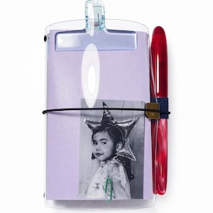 May include: A clear clipboard with a light purple notepad, a black and white photo, and a red pen. The photo is of a child wearing a star-shaped headpiece. A green paper clip secures the photo to the notepad.