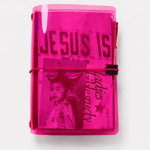 May include: A bright pink translucent plastic notebook cover featuring a black elastic band closure. The cover displays a photograph of a person wearing a star-shaped headpiece, with text that reads JESUS IS, ladies bible study, and 2ND & 4TH W.