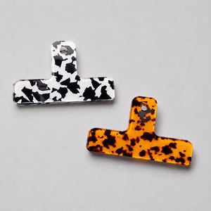 May include: Two T-shaped clips with a flat, rectangular base and a vertical tab featuring a circular hole. One clip has a black and white cow-print pattern, while the other features a translucent amber and black tortoiseshell pattern.