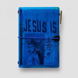 May include: A rectangular notebook with a translucent blue plastic cover held shut by a thin blue elastic band. The cover displays the text "JESUS IS" in bold letters above a photograph of a person wearing a star-shaped hat and the word "ladies".
