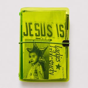 May include: A neon yellow translucent notebook cover featuring the text "JESUS IS" at the top. The front insert displays a black and white photograph of a person wearing a star-shaped headpiece, alongside the text "ladies bible study 2nd & 4th w".