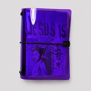Può includere: Un raccoglitore in plastica viola traslucido con chiusura elastica nera. La copertina mostra il testo "JESUS IS" e "Ladies Bible Study 2nd & 4th W" accanto a una fotografia in bianco e nero di una persona che indossa un copricapo a stella.