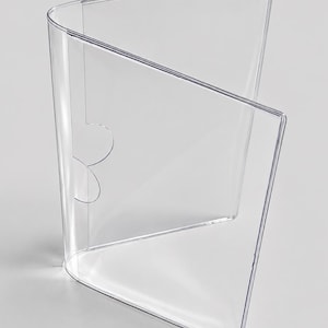 Könnte beinhalten: Ein transparenter, dreiteiliger Menühalter oder Aufsteller aus Acryl. Die Struktur besteht aus drei vertikalen Paneelen, die durch einen gebogenen Rücken verbunden sind, mit einem halbkreisförmigen Ausschnitt an der Seite zum Einlegen.