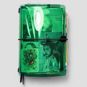 May include: A transparent green planner with a black elastic closure. Inside are various images and text that reads "2nd & 4th Weeks Of Tuesday Fridays". A gold pen is attached to the side. The planner is on a white background.