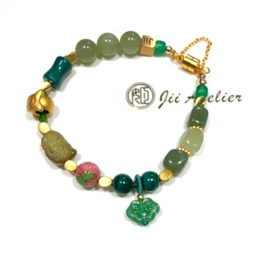 May include: A beaded bracelet featuring a variety of green and gold beads, including round, cylindrical, and irregular shapes. The bracelet has a gold clasp and a green charm. The text "Jii Atelier" is visible.