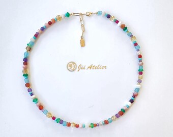 May include: A colorful beaded necklace with a gold clasp and extender chain. The necklace features a variety of small, irregularly shaped beads in shades of blue, green, red, yellow, purple, and white. The text "Jii Atelier" is visible.