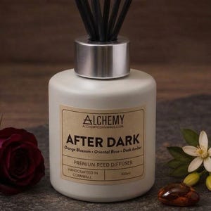 May include: A matte white reed diffuser bottle with a silver cap and black reeds. The label reads Alchemy After Dark, Orange Blossom, Oriental Rose, Dark Amber, Premium Reed Diffuser, Handcrafted in Cornwall, 100ml. A dark red rose and white flower sit nearby.