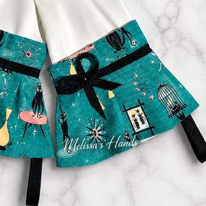 May include: A pair of white sleeve cuffs featuring a teal fabric overlay with a mid-century pattern of black cats, birdcages, and starbursts. Each cuff is accented with a black ribbon bow and a black ribbon loop at the bottom. The text Melissa's Hands is printed in white script on the lower right side.